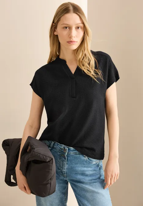 Frau mit langem Haar trägt schwarzes Shirt und Jeans, hält eine Jacke in der linken Hand.